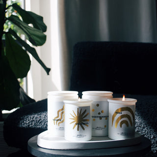 four lit candles in white jars in a room with a black couch and the sun coming in through the window