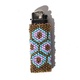 A clear plastic lighter covered with a handmade beaded sleeve featuring a geometric pattern of lavender, light blue, and metallic gold seed beads, shown on a white background with a soft shadow.