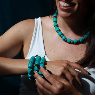 A woman wearing a chunky turquoise beaded necklace and matching beaded bracelets, shown in a close-up lifestyle shot against a dark background.