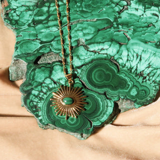 A shiny 14k  gold fill necklace in the shape of a sunburst pendant with a malachite accent in the middle, laying on a 14k gold fill malachite slab.