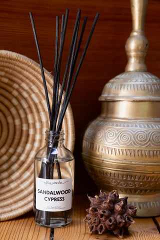 A clear glass reed diffuser bottle labeled 'SANDALWOOD CYPRESS' with several black reeds, styled on a wooden surface beside a woven tray, a brass decorative vessel, and a brown seed pod.