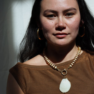 Woman wearing a brown top and necklace with a shell pendant against a neutral background