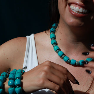 A close-up lifestyle shot of a woman wearing a turquoise beaded necklace and matching beaded bracelets against a dark background.