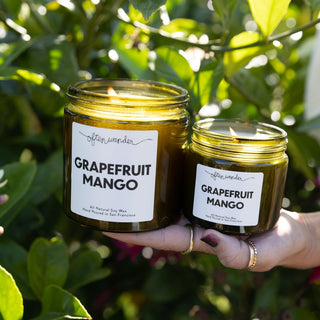 Two amber glass jar candles labeled 'Grapefruit Mango' by often wander, held in a person's hands against a background of green citrus leaves.