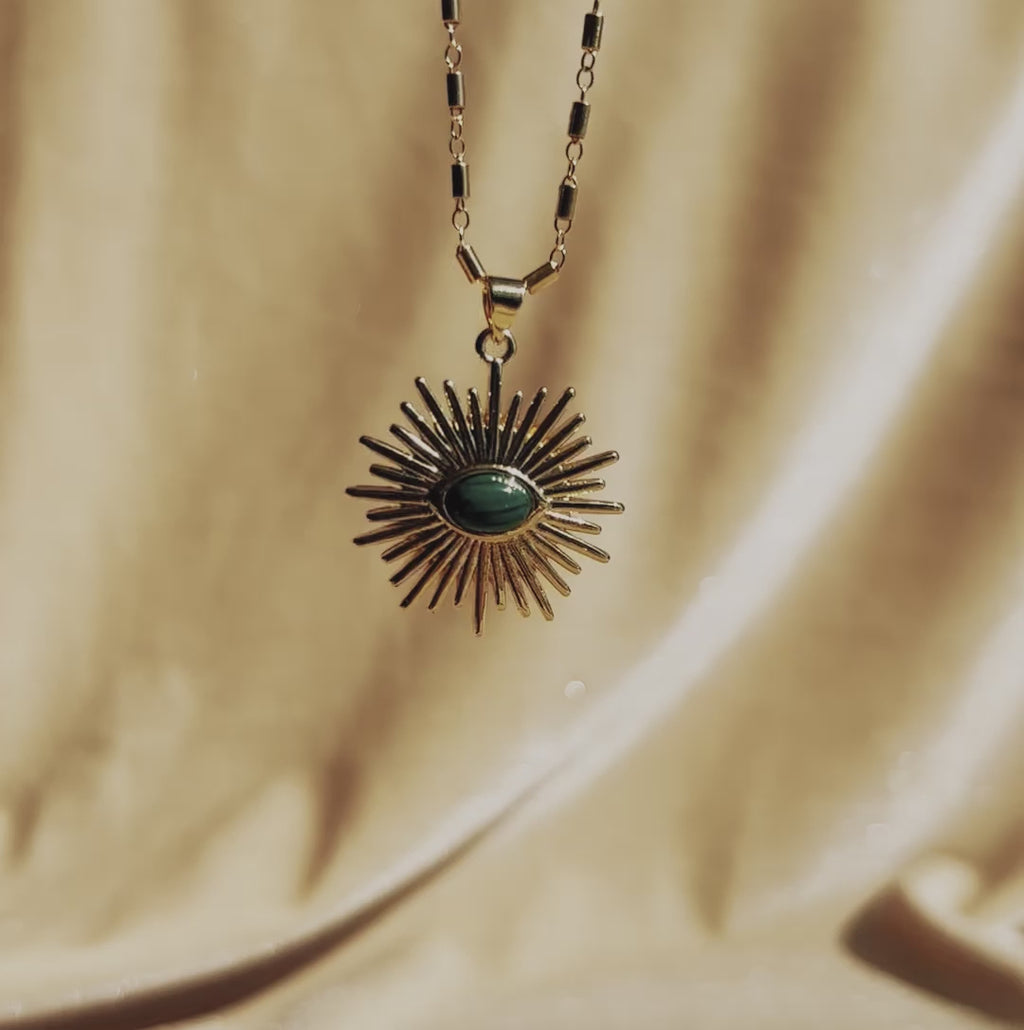 A shiny 14k gold fill necklace in the shape of a sunburst pendant with a malachite accent in the middle, laying on a 14k gold fill malachite slab.